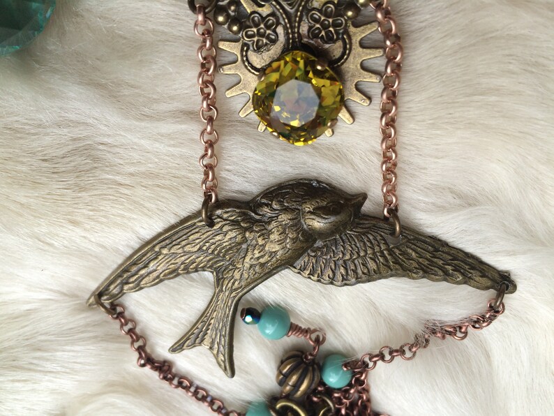 May include: A vintage-style necklace featuring a bronze-colored bird pendant with outstretched wings. The necklace includes a large, faceted yellow-green gemstone, decorative metalwork, and copper-colored chain accents. Small turquoise beads add a pop of color.