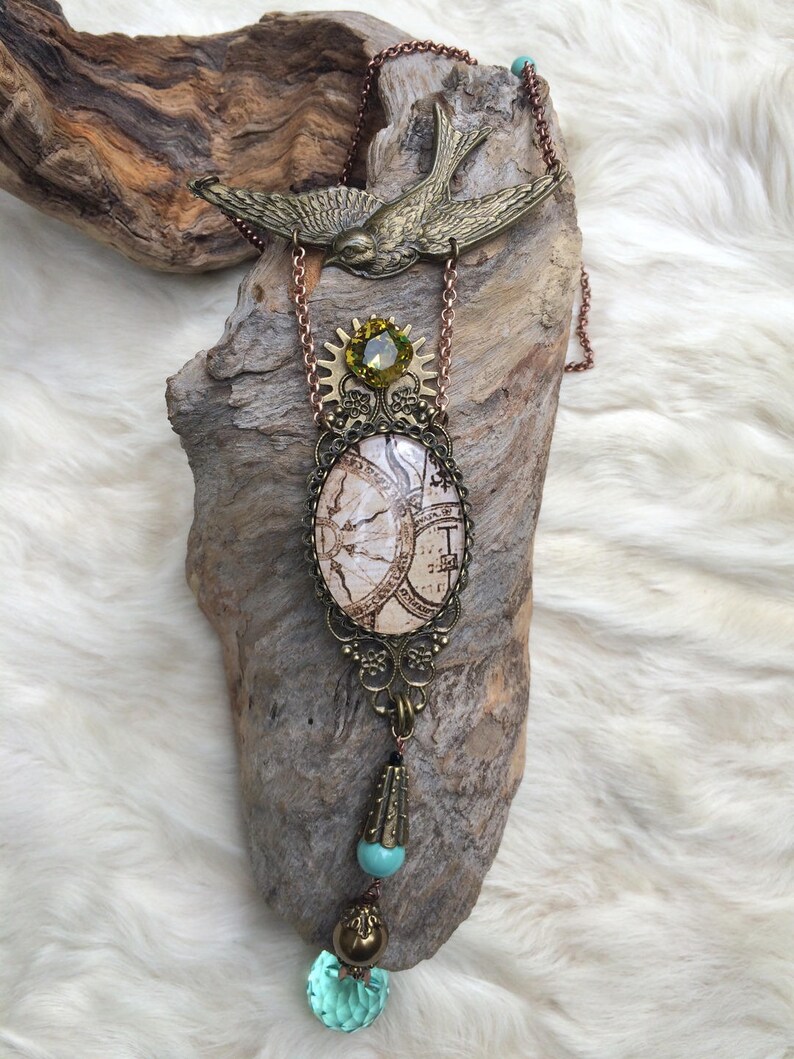 May include: A steampunk-style necklace featuring a brass bird pendant and a large oval pendant with a map design. The necklace includes a green gemstone, turquoise beads, and a faceted aqua crystal. The chain is a copper color.