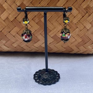 May include: A pair of dangle earrings featuring a dark red glass bead, gold accents, and colourful enamel details. The earrings are displayed on a black stand.