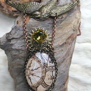 May include: A steampunk-style necklace featuring a bronze-colored bird pendant at the top. Below, a green gemstone sits atop a gear-shaped element, connected to an oval pendant with a vintage map design. The chain is a copper color.