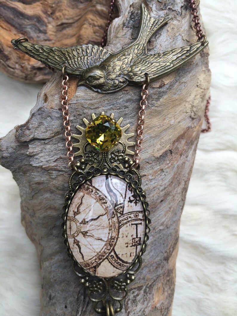 May include: A steampunk-style necklace featuring a bronze-colored bird pendant at the top. Below, a green gemstone sits atop a gear-shaped element, connected to an oval pendant with a vintage map design. The chain is a copper color.