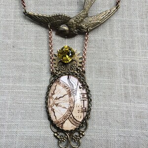 May include: A vintage-style necklace featuring a brass-toned swallow pendant at the top. Below, a yellow gemstone sits above an oval pendant with a map design. The necklace has a copper-colored chain and decorative filigree.