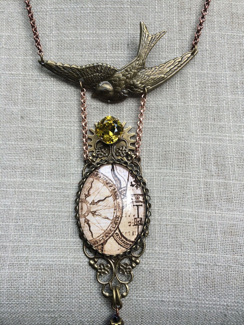 May include: A vintage-style necklace featuring a brass-toned swallow pendant at the top. Below, a yellow gemstone sits above an oval pendant with a map design. The necklace has a copper-colored chain and decorative filigree.