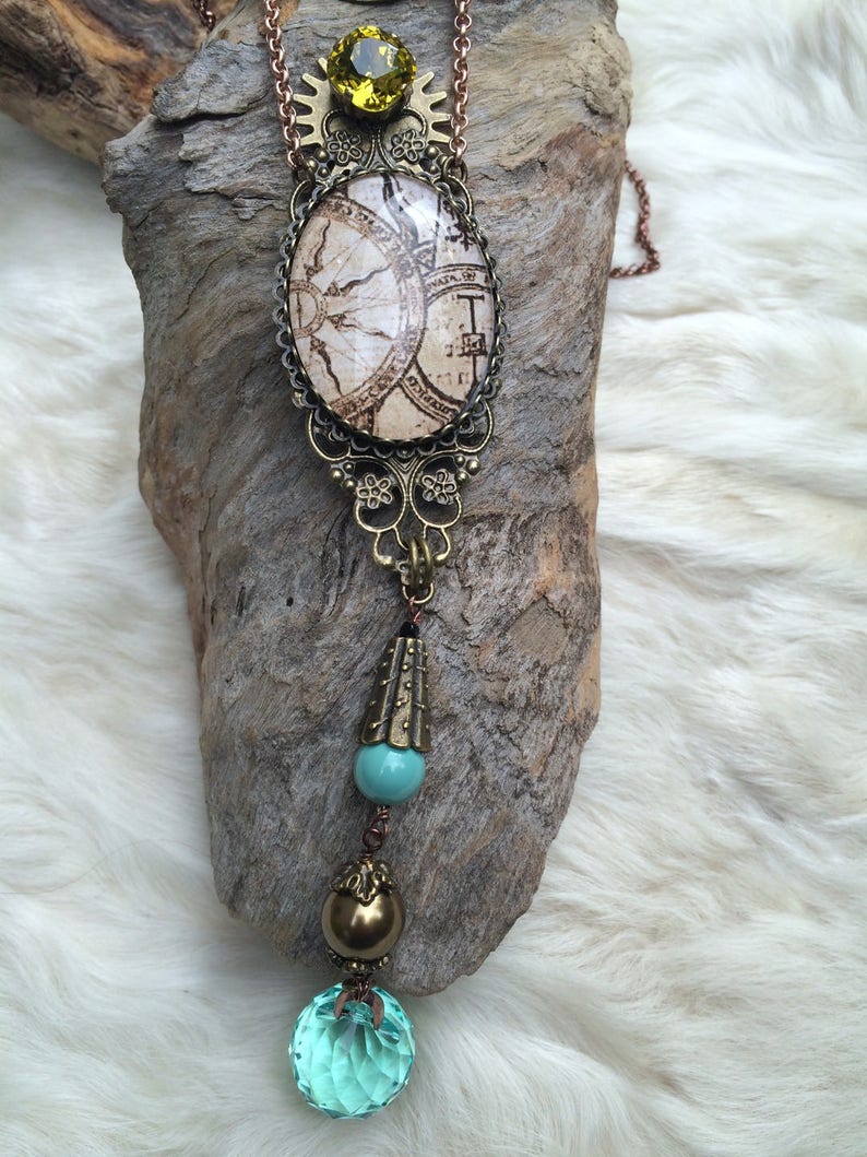 May include: A steampunk-style necklace with a vintage map design in an oval pendant. The necklace features a large, faceted, light blue crystal at the bottom, with brass and turquoise accents. A large, yellow-green gemstone sits at the top.