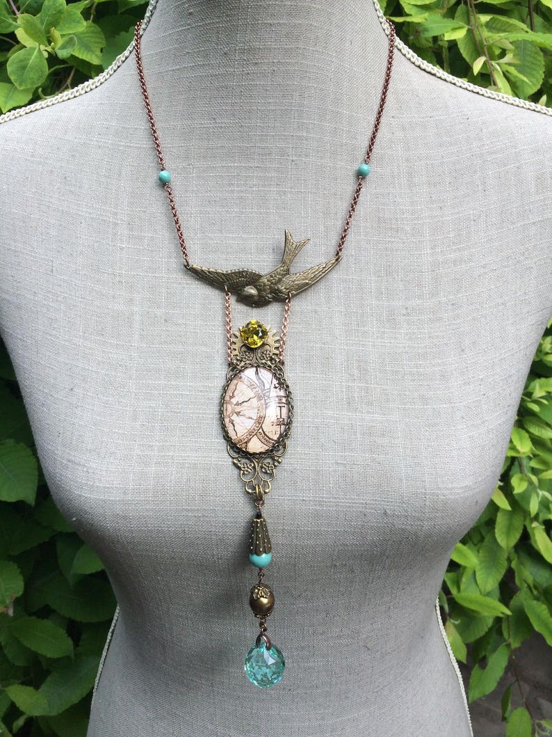 May include: A vintage-style necklace with a bronze-colored chain and pendant. The pendant features a bird, a clock face design, and various beads in shades of turquoise, gold, and a clear blue. The necklace is displayed on a mannequin.