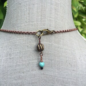 May include: Copper-colored chain necklace with a turquoise and black bead pendant. The necklace features a lobster clasp and a decorative bronze-colored bead. The pendant is suspended from a copper wire.