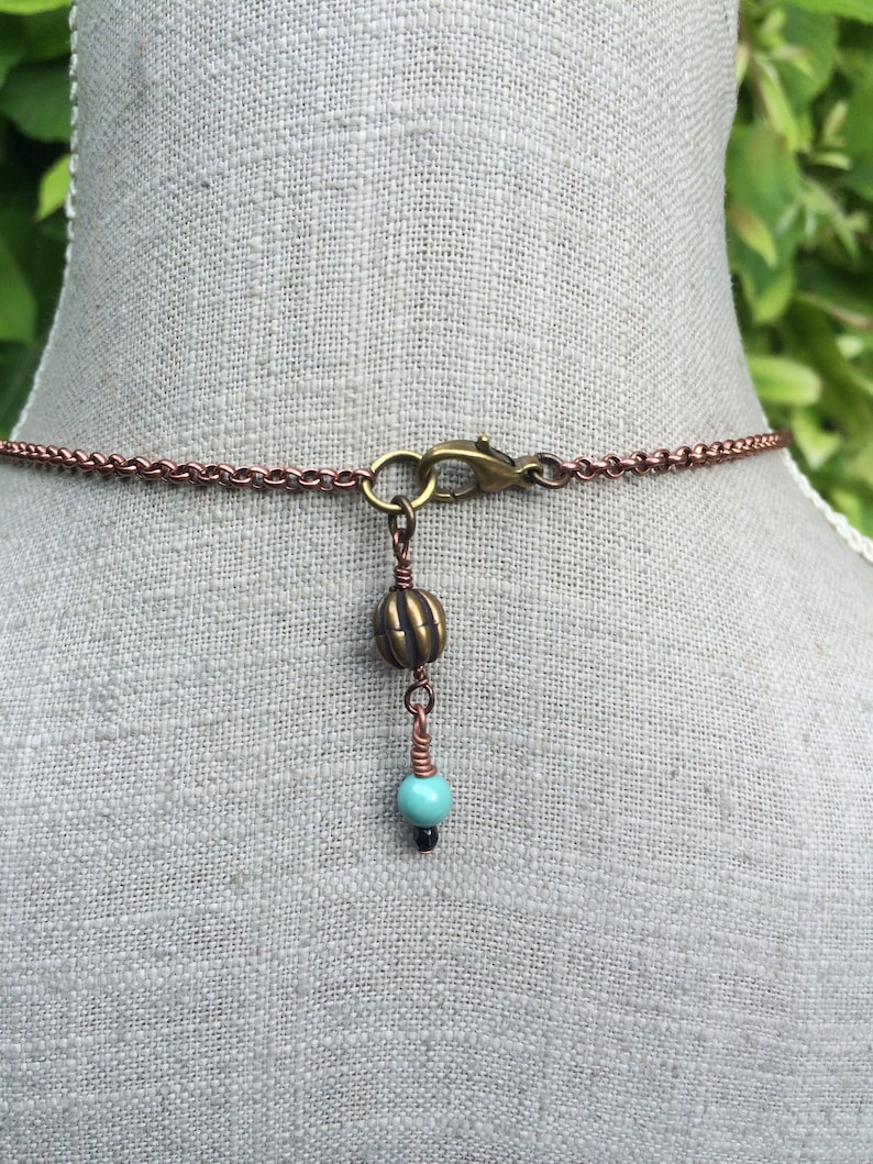 May include: Copper-colored chain necklace with a turquoise and black bead pendant. The necklace features a lobster clasp and a decorative bronze-colored bead. The pendant is suspended from a copper wire.