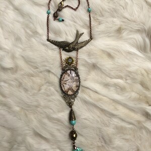 May include: A long, antique-style necklace with a bronze chain and pendant. The pendant features a bird, an oval-shaped design, and a faceted, light blue crystal. The necklace has turquoise-colored beads and a decorative clasp.