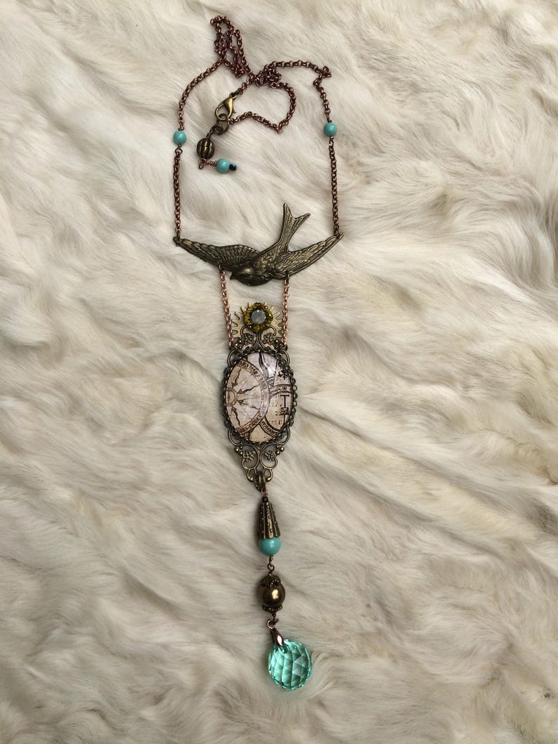 May include: A long, antique-style necklace with a bronze chain and pendant. The pendant features a bird, an oval-shaped design, and a faceted, light blue crystal. The necklace has turquoise-colored beads and a decorative clasp.