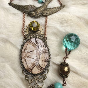 May include: A vintage-style necklace featuring a bronze-toned swallow pendant and an oval pendant with a map design. The necklace includes a yellow gemstone, turquoise beads, and a faceted green crystal. The chain is a copper color.