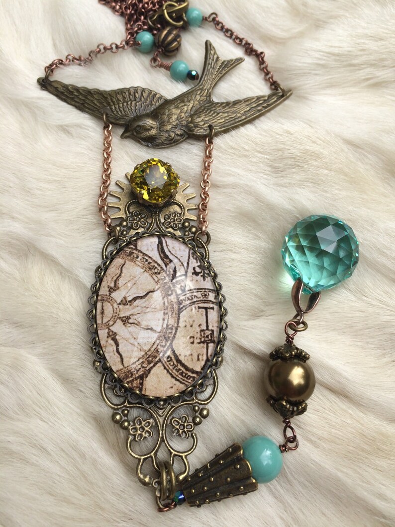 May include: A vintage-style necklace featuring a bronze-toned swallow pendant and an oval pendant with a map design. The necklace includes a yellow gemstone, turquoise beads, and a faceted green crystal. The chain is a copper color.