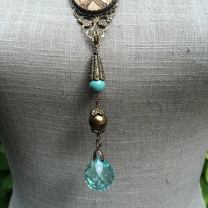 May include: A vintage-style pendant necklace with a map design. The necklace features a large, round pendant with a map design, intricate filigree, a turquoise bead, a gold bead, and a faceted, light blue crystal. The necklace is displayed on a mannequin.