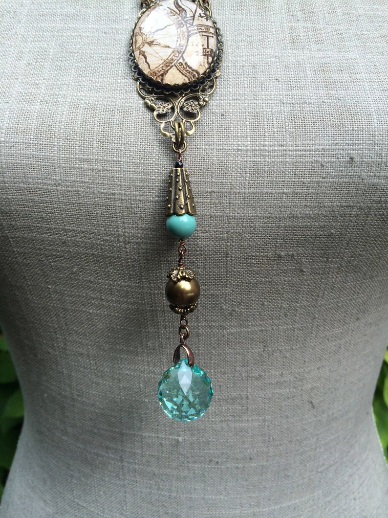 May include: A vintage-style pendant necklace with a map design. The necklace features a large, round pendant with a map design, intricate filigree, a turquoise bead, a gold bead, and a faceted, light blue crystal. The necklace is displayed on a mannequin.