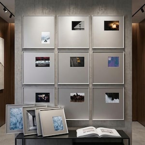 Pode incluir: Uma parede de galeria com nove molduras quadradas grandes com bordas prateadas finas, cada uma contendo uma pequena fotografia centralizada. Três molduras adicionais combinando estão apoiadas numa mesa consola preta, junto a um livro aberto.