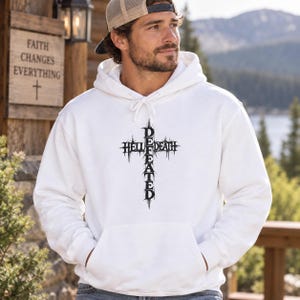 Puede incluir: Una sudadera con capucha blanca que presenta un diseño gráfico negro de una cruz formada por las palabras HELL y DEATH que se cruzan con la palabra DEFEATED. La sudadera incluye un bolsillo tipo canguro y cordones blancos en el cuello.