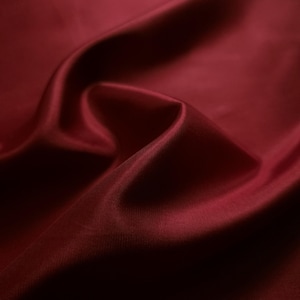 May include: A deep burgundy fabric with a smooth, slightly reflective surface. The material is draped in soft, flowing folds that create subtle highlights and shadows, emphasizing the rich, dark red color and the fine, uniform texture of the weave.