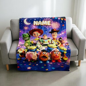 May include: A rectangular fleece blanket featuring a vibrant night sky background with fireworks, stars, and the word NAME at the top. The design includes a group of animated characters including a cowboy, space ranger, dinosaur, and potato figures.