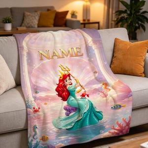May include: A soft throw blanket featuring a colorful illustration of a red-haired character with a golden crown and trident, sitting on a purple cushion inside a large seashell. The text NAME is printed in gold above the character with sea life.