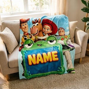 Puede incluir: Una manta de forro polar azul claro con los personajes de Toy Story: Woody, Bullseye, Jessie y Buzz Lightyear. Un gran marco verde con forma de rana está centrado en la parte inferior con la palabra NAME impresa en letras amarillas.