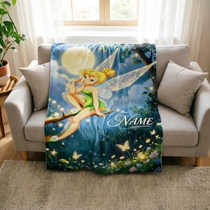 May include: A soft throw blanket featuring a printed illustration of a blonde fairy with translucent wings sitting on a tree branch against a night sky with a large moon. The design includes glowing butterflies, flowers, and the word NAME in script.