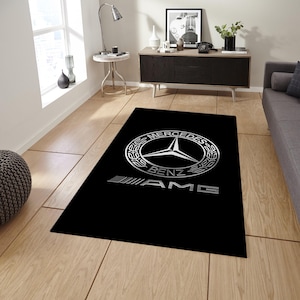 Könnte beinhalten: Ein rechteckiger schwarzer Teppich mit dem silbernen Mercedes-Benz-Logo, das oben zentriert ist und von einem kreisförmigen Lorbeerkranz umschlossen wird, wobei sich der silberne AMG-Schriftzug direkt unter dem kreisförmigen Emblem befindet.