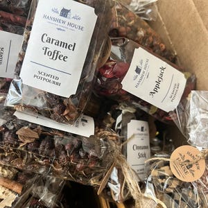 May include: Multiple clear plastic bags filled with scented potpourri, featuring dried botanicals, wood pieces, and fabric accents. Labels display the Hanshew House logo and scent names including Caramel Toffee, Applejack, and Cinnamon Stick.