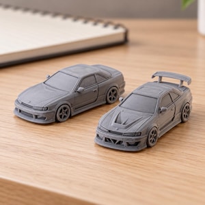 May include: Two small gray model cars made of a matte material. The car on the left is a coupe with a smooth hood. The car on the right is a coupe featuring a vented hood and a rear spoiler. Both vehicles have detailed wheels and body panels.