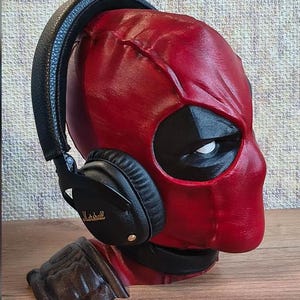 以下が含まれることがあります： 黒いマーシャル製ヘッドホンを装着した、赤と黒のデッドプールのキャラクターバスト。バストは質感のある赤いマスクに黒い目の周りのパーツ、暗い色の肩当てが特徴です。ヘッドホンは黒いヘッドバンドとイヤーカップを備えています。