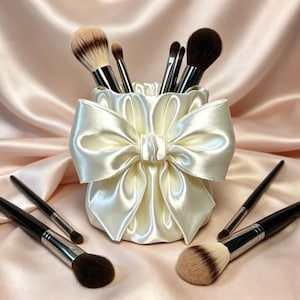 Puede incluir: Un porta brochas de maquillaje de satén color crema con un gran lazo plisado en la parte frontal. El recipiente cilíndrico contiene varias brochas de maquillaje con mangos negros y cerdas marrones, mientras otras brochas yacen al lado.