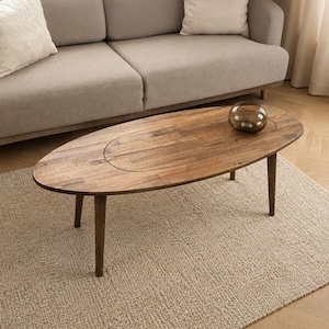 May include: An oval wooden coffee table with a natural grain finish and four tapered legs. The tabletop features a distinct central oval inlay panel. A small, round, dark-tinted glass decorative bowl sits on the right side of the table surface.