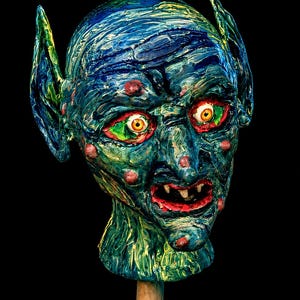 May include: A sculpted head of a creature with pointed ears, textured blue and green skin, and small red bumps. The face features wide, yellow eyes with red surrounding irises, a wide mouth with visible teeth, and a thick, layered paint finish.