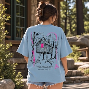 May include: A light blue t-shirt featuring a black ink illustration of a cabin on chicken legs in a forest under a moon. The cabin window is bright pink. Text reads: WOMEN? OH, WE'RE ANGRY ABOUT EVERYTHING. ENCORE NATION. The design includes pink accents.