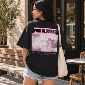 Peut inclure: Un t-shirt noir à manches courtes avec un graphique rectangulaire de nuages roses. Le texte PINK CLOUDING apparaît en lettres noires grasses sur un fond rose au-dessus des nuages, avec le texte but I was pink clouding for the thrill of it et ENCORE NATION imprimé en dessous.