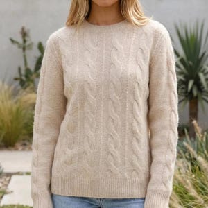 Può includere: Un maglione beige a trecce con scollo rotondo e maniche lunghe, caratterizzato da un motivo a trecce verticali testurizzate sul davanti e polsini e orlo a coste, abbinato a un paio di jeans azzurri.