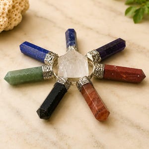May include: A star-shaped crystal grid featuring six pointed gemstone wands in blue, green, black, orange, red, and dark purple, each capped with a silver-toned metal bail, surrounding a central clear, hexagonal, flat-topped quartz crystal point.