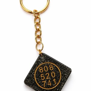 May include: A diamond-shaped keychain featuring a black glittery surface with a gold-colored circular inlay. The inlay displays the numbers 808, 520, and 741 in a gold font. The charm is attached to a gold-colored metal split ring by a short chain.