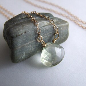 May include: A delicate gold chain necklace with a light green gemstone pendant. The pendant is faceted and has a teardrop shape.