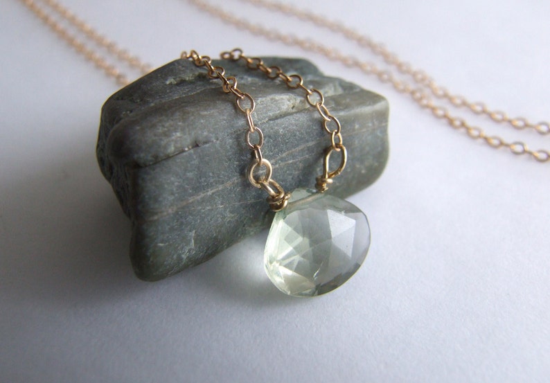 May include: A delicate gold chain necklace with a light green gemstone pendant. The pendant is faceted and has a teardrop shape.