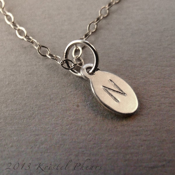 May include: A silver-toned necklace with a small, oval pendant. The pendant features the letter "N" engraved on its surface. The chain is delicate, and the pendant is attached with a small ring. The background is a neutral gray.
