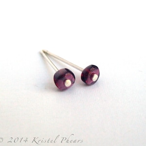 May include: A pair of small, silver stud earrings with round, dark purple glass beads. The beads have a white center.