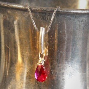 May include: A silver necklace with a teardrop-shaped, faceted red gemstone pendant. The pendant is wire-wrapped and hangs from a silver bar and chain. The jewelry is displayed against a metallic background.