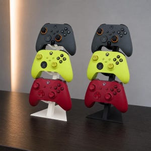 Könnte beinhalten: Zwei vertikale Gaming-Controller-Ständer, einer in Weiß und einer in Schwarz, die jeweils drei kabellose Controller in Dunkelgrau, Neongelb und Dunkelrot halten. Die Controller verfügen über Standard-Tastenlayouts, Analogsticks und das Xbox-Logo.