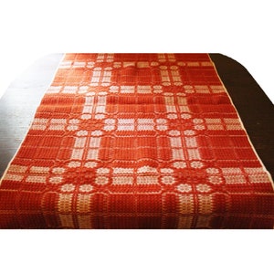May include: A red and white woven table runner with a geometric pattern. The runner is made of a thick, textured fabric and has a fringed edge.