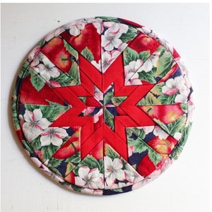 May include: Round red, white and green fabric hot pad with a star design. The star is made of red fabric and the background is a floral print.