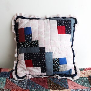 May include: Square patchwork cushion with a light pink centre and a ruffled edge. The patchwork design features squares of red, blue, and navy fabrics. The cushion is approximately 40 cm square.