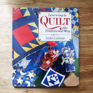 May include: A book cover for "Learning to Quilt the Traditional Way" by Annlee Landman. The design includes a patchwork quilt in red, blue, and yellow. Two fabric puppets and a small tote bag are also visible.