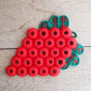 May include: A handmade, knitted trivet shaped like a bunch of red grapes. The grapes are made of small, round, red yarn circles. The leaves are green with red detailing. The trivet rests on a wooden surface.