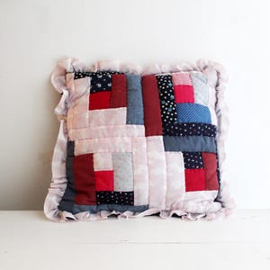 May include: Square patchwork cushion with a frilled edge. The cushion features fabric squares in red, navy, light blue, grey, and white. The frilled edge is a light pink with a subtle pattern.