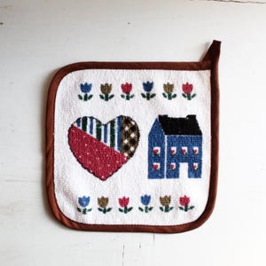 May include: White quilted pot holder with a brown border. The design features a patchwork heart, a blue house, and rows of embroidered flowers in blue, brown, and red. A loop is attached for hanging.