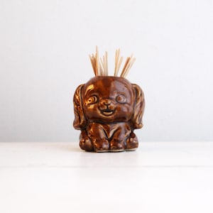 May include: A brown ceramic dog-shaped toothpick holder. The holder is glossy and features a smiling face and floppy ears. Several wooden toothpicks are inserted in the top of the dog's head. The holder is sitting on a white surface.